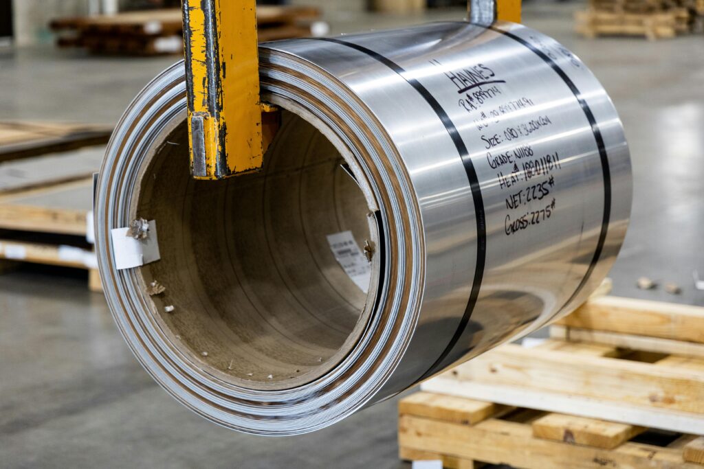 "Stainless steel coil roll suspended by industrial lifting equipment in a warehouse, showing product details like grade, size, and weight for metal supply or fabrication use."